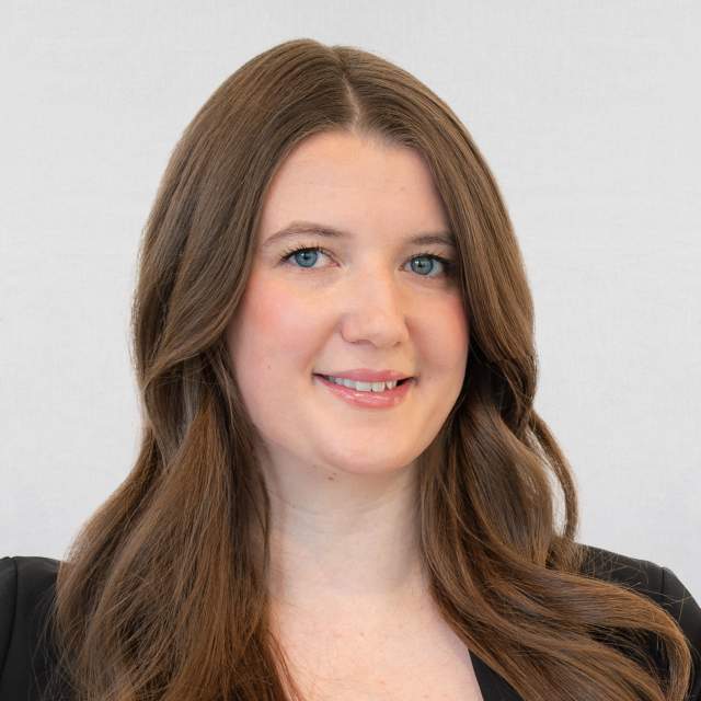Professional headshot of a woman with long brown hair wearing a black top, smiling slightly against a light gray background.