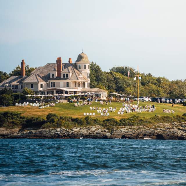 A large white building with a gray roof sits on a grassy hill overlooking the ocean, with people and white chairs on the lawn.