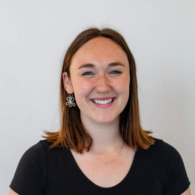 A young woman with straight, shoulder-length brown hair smiles at the camera. She is wearing a black short-sleeve top, dangling flower-shaped earrings, a small nose ring, and a delicate necklace. The background is plain and light-colored.