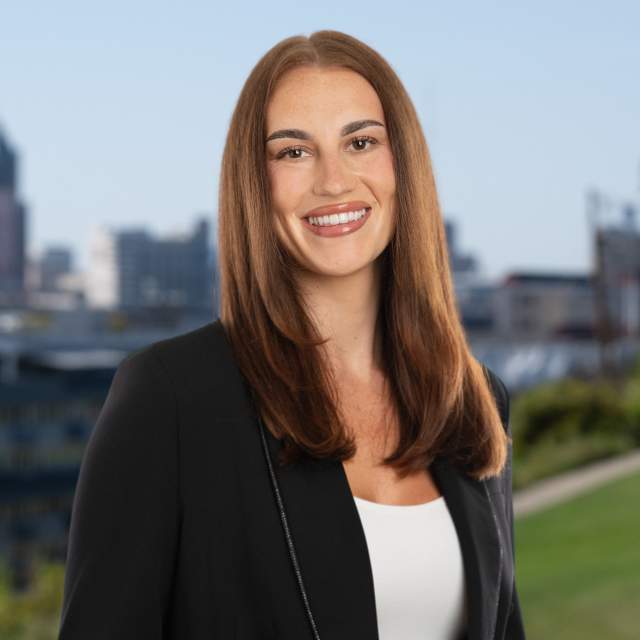 Professional headshot of a smiling Abi Hammonds in a black blazer standing outdoors, with Milwaukee's skyline softly visible in the background.