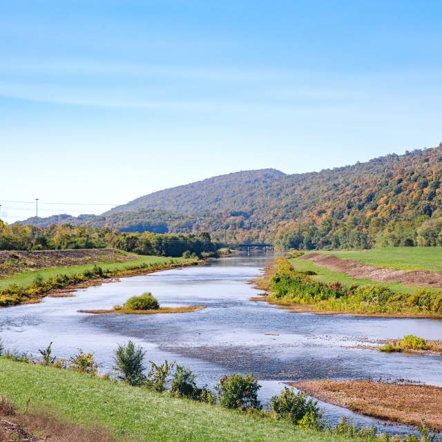 River winding through a grassy valley with tree-covered mountains in the background under a clear blue sky.
