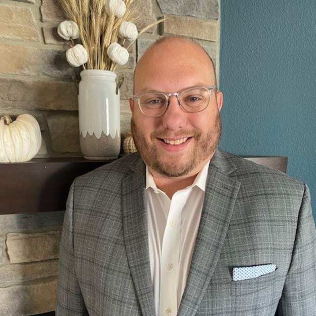 Ben Risseeuw in glasses smiles at the camera, wearing a gray plaid blazer over a white shirt, standing indoors near a stone wall with neutral décor.