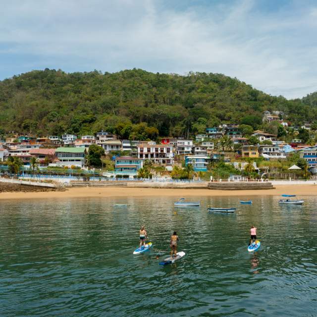Tres personas practican paddleboard en aguas tranquilas cerca de la playa y casas coloridas en la ladera de Isla Taboga, rodeadas de exuberante vegetación.