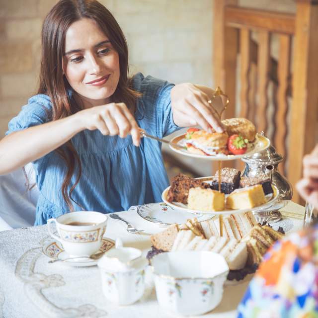 A woman in a blue dress takes a scone from a cake stand, which is filled with sandwiches, scones and cakes, in the Cotswolds