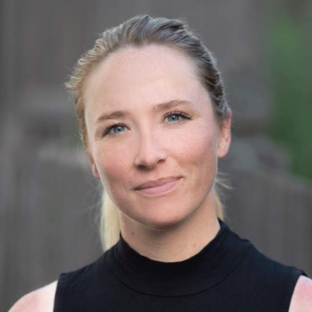 Portrait of a woman with blonde hair tied back, wearing a sleeveless black top, looking at the camera with a neutral expression. The background is softly blurred.