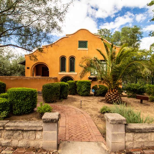 A small yellow house with a unique style of Southwestern architecture peaking out from behind its green desert lawn