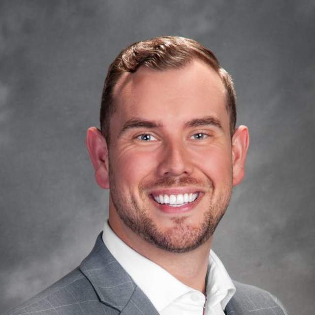 Professional portrait of a smiling man in a grey checkered suit jacket and white dress shirt, set against a grey studio background.
