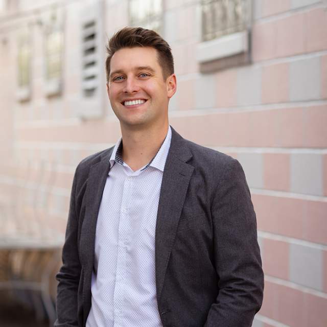 A white man wearing a dark grey suit coat and white button up, smiling with his hands in his pockets.