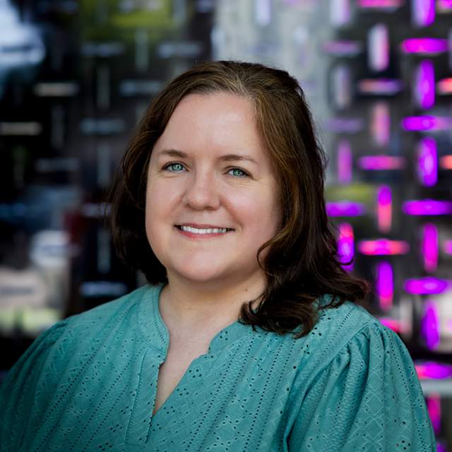 A woman with brown hair wearing a teal blouse, standing in front of a wall with bright purple lights.