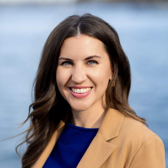 A woman in a beige blazer and a blue blouse underneath standing in front of the lake.