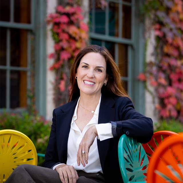 A white women in a white button down and navy blazer sitting on the sunburst chairs at the Memorial Union Terrace.