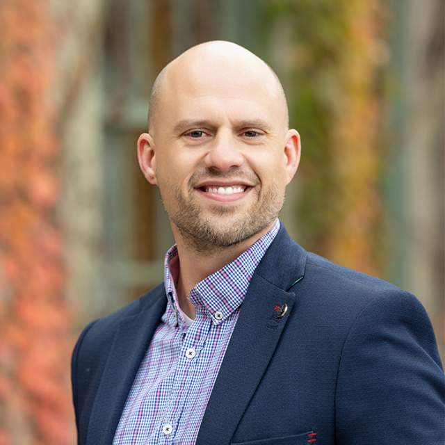 A white man wearing a plaid button up under a dark blue blazer standing in front of a wall full of foliage.
