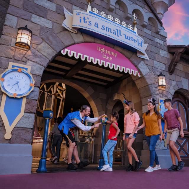 A family entering the "It's a Small World" attraction at a Disney theme park, greeted by a cast member, with a colorful sunset in the background.