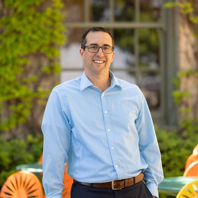 A white man with glasses wearing a light blue button down shirt standing around color terrace chairs.