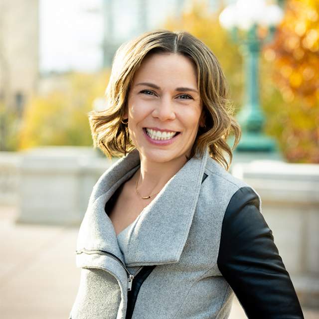 A woman in a grey jacket with black leather sleeves smiles and stands in front of fall colored trees.