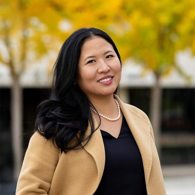 A woman wearing a black shirt under a tan blazer standing in front of fall colored trees.