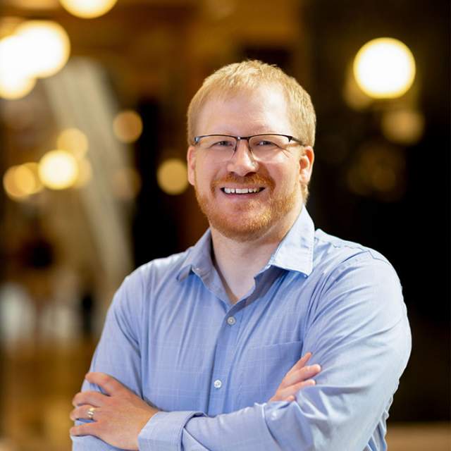 A man with orange hair and glasses wearing a blue button up shirt standing his with his arms crossed.