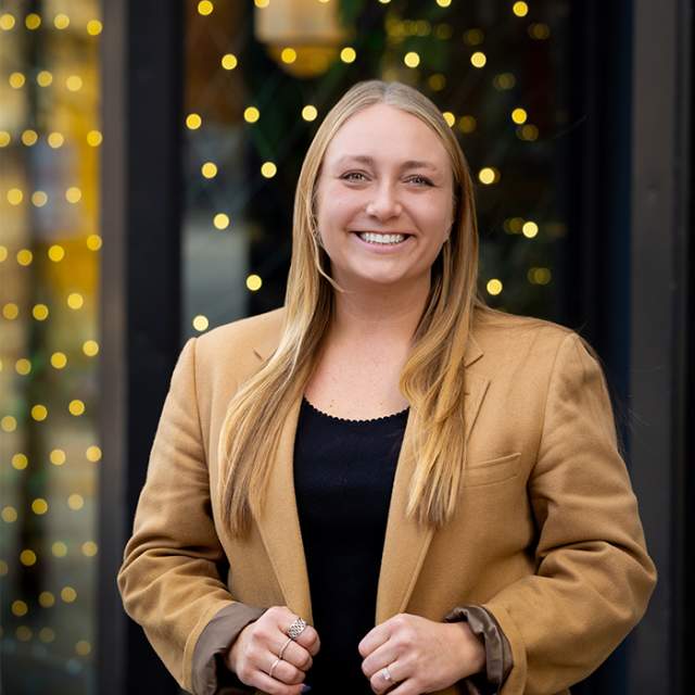 A white woman standing in front of a building with holiday lights hanging in front of the windows.