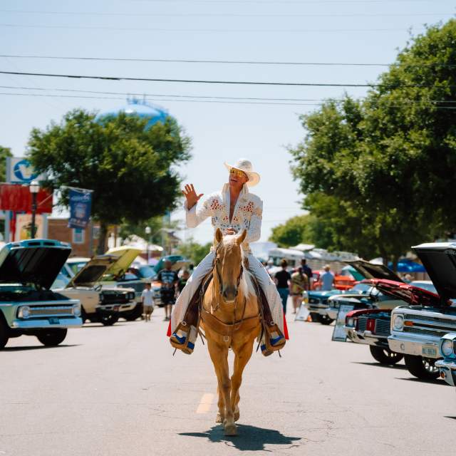 ChatGPT said:

A man dressed as Elvis in a rhinestone-studded white jumpsuit and cowboy hat rides a palomino horse down a street lined with classic cars at a Route 66 festival finale event in Amarillo.