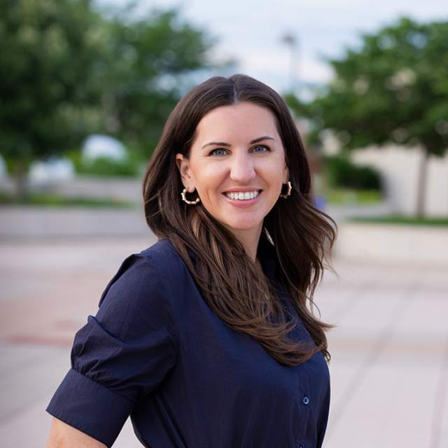 A woman with dark hair wearing a navy button up dress, standing outside with trees behind her.