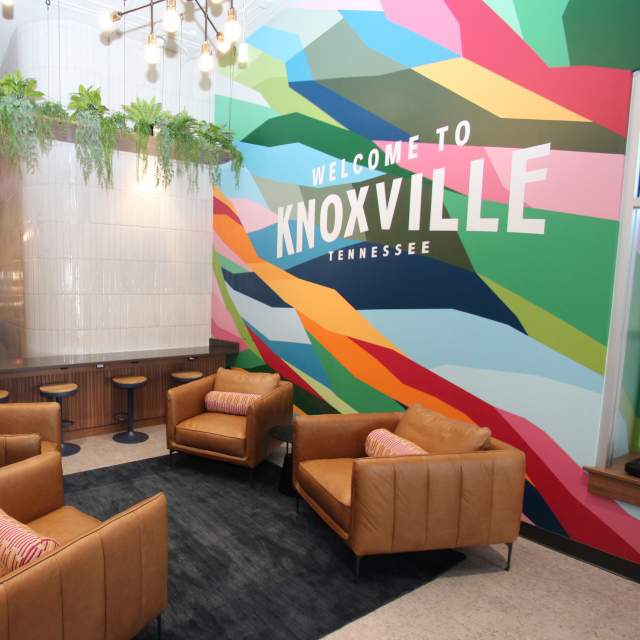 The modern interior of the Visit Knoxville Visitors Center featuring a lounge area and a vibrant mural. In the foreground, four tan leather armchairs with colorful patterned pillows are arranged around a dark area rug. The background is dominated by a large, floor-to-ceiling mural composed of bright geometric shapes in shades of blue, green, pink, and orange, with the words "WELCOME TO KNOXVILLE TENNESSEE" printed in bold white letters. To the left, a wood-paneled counter with blue barstools sits beneath a white tiled pillar and a wooden ceiling fixture overflowing with hanging green plants. Exposed bulb light fixtures hang from the ceiling, adding to the contemporary industrial feel.