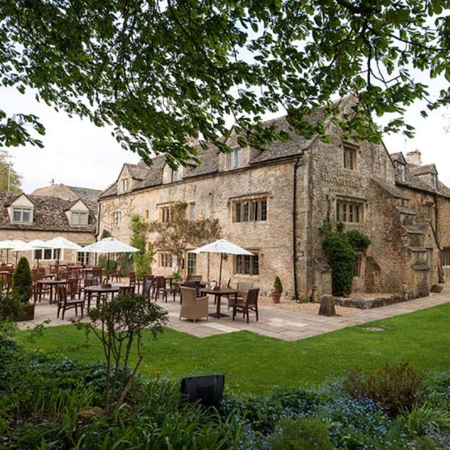Wooden tables and chairs with white parasols sit on a patio behind The Slaughters Country Inn, surrounded by gardens