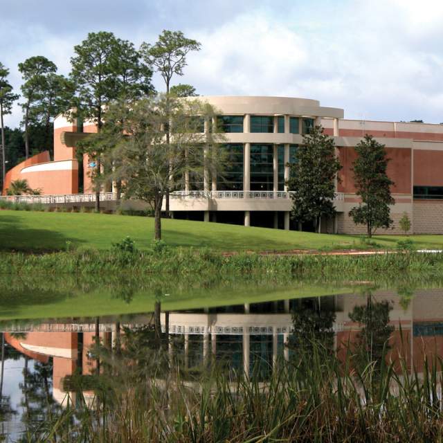 The Mobile Museum of Art building with curved architecture reflects in a serene pond, surrounded by lush green grass and trees.