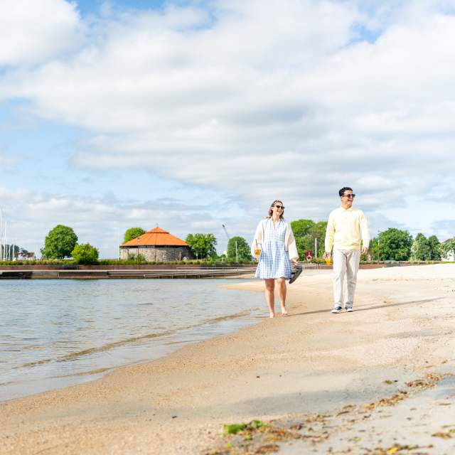 En mann og en kvinne går på tur langs vannkanten på Bystranda i Kristiansand i solskinn