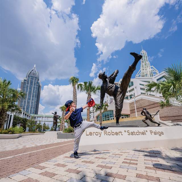 A young boy in a baseball uniform emulates the pitch of a statue behind him