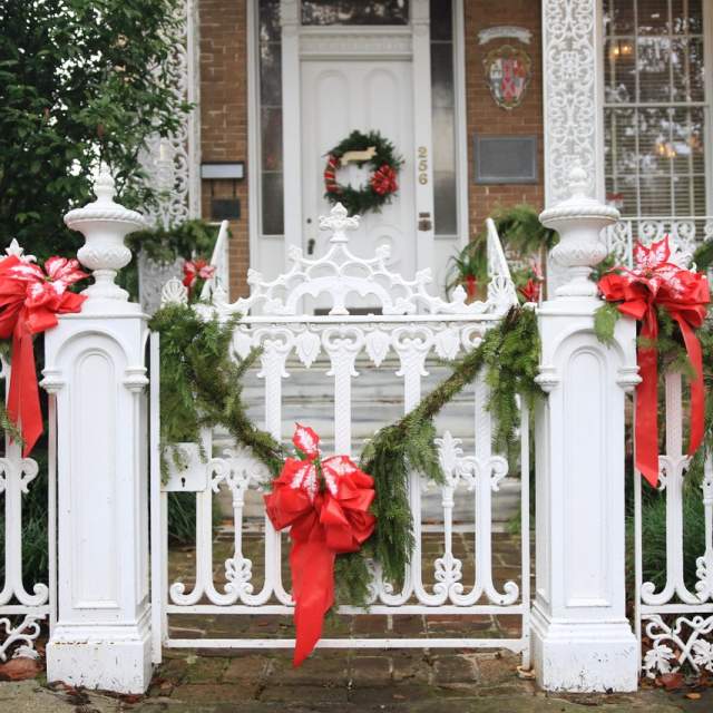 White cast iron fence with green leafy garland and red bows in front of a historic brick home