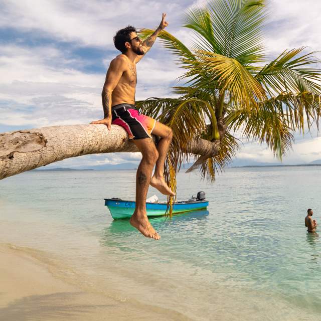 Homem sentado numa palmeira numa praia caribenha com um oceano azul claro cristalino e pessoas ao fundo.