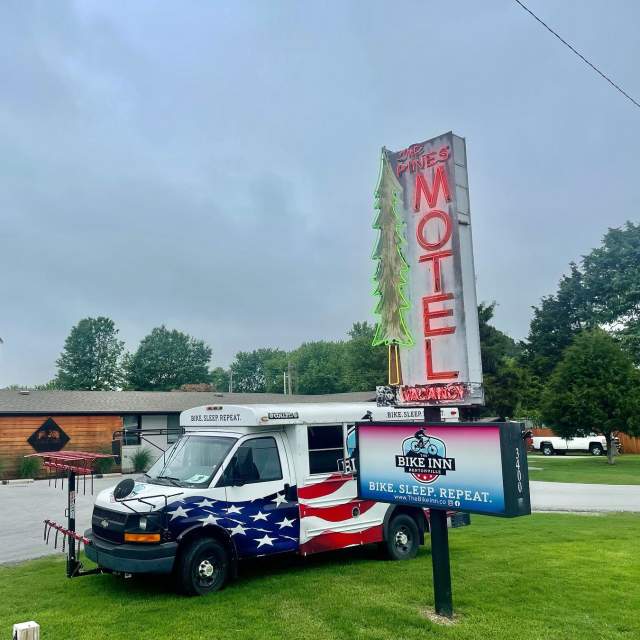 American flag-themed shuttle bus in front of 'BIKE INN' sign with a neon-lit 'PINES MOTEL' sign overhead on an overcast day.