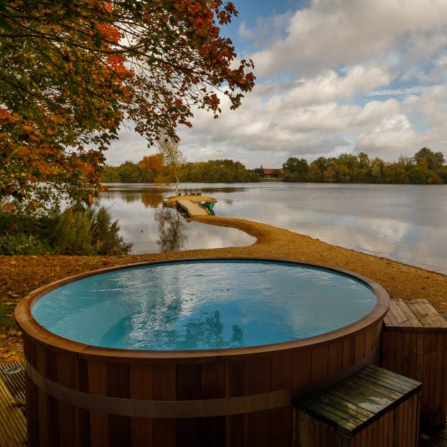 A wooden hot tub on a deck with views across the lake and leaves falling from the trees, at Log House Holidays in the Cotswolds