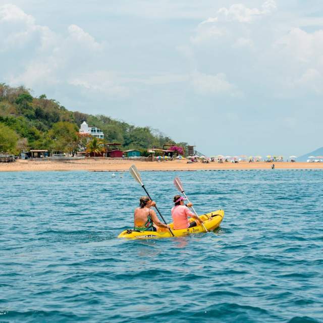 Duas pessoas remam num caiaque amarelo nas águas azuis perto de uma praia de areia clara e uma ilha verde e montanhosa sob um céu parcialmente nublado.
