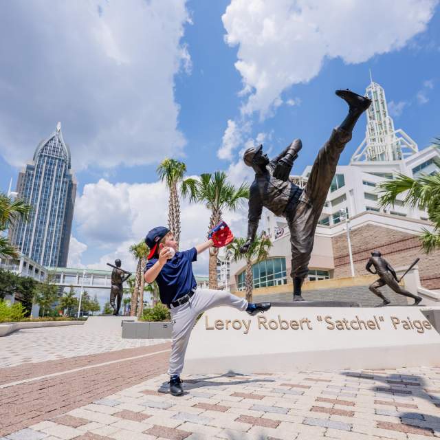 a young boy in baseball gear posed as if to throw a baseball next to an iron statue of baseball player Leroy Robert "Satchel" Paige at the Hall of Fame Walk Plaza. The Mobile Convention Center and Renaissance Riverview Plaza Hotel are seen in the skyline.