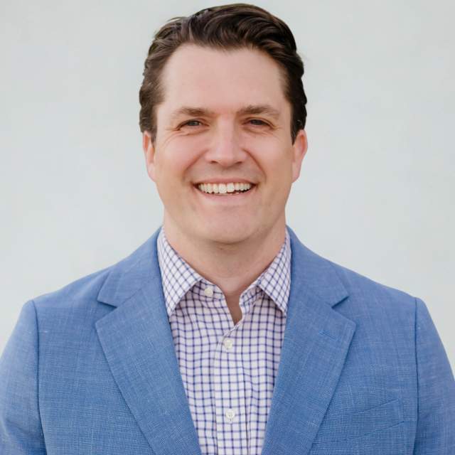 smiles against a clean, neutral background, wearing a light blue blazer over a patterned button-down shirt for a polished, professional portrait.