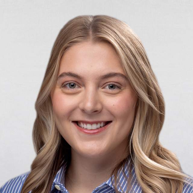 Professional headshot of a smiling woman with long, wavy blonde hair wearing a blue and white striped button-down shirt, posed against a light gray background.