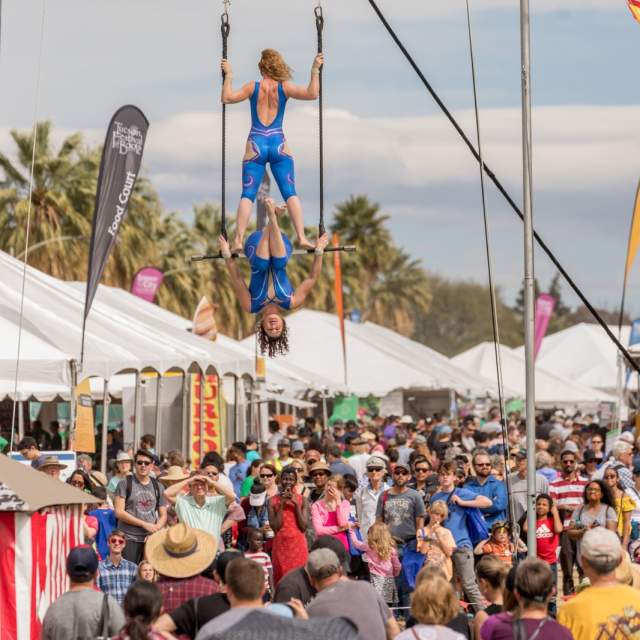 Large crowd outdoors between white tent booths. Two acrobats on a bar hang over the crowd