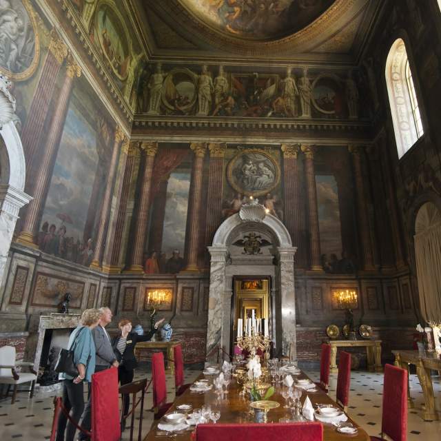 A couple with a tour guide stand in an impressive room with painted images on the ceiling and walls, large marble doorways and gold furniture at Blenheim Palace