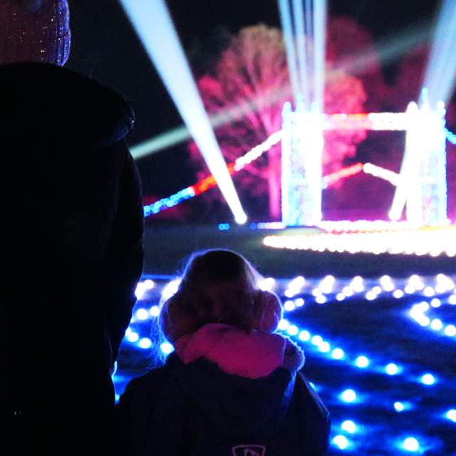 A mother and small child stand with their backs to the camera looking at a small, lit up London Bridge. There are roaming spotlights & glowing fairylights on the floor in all different colours. The trees off in the distance are also lit up in blue and pink.