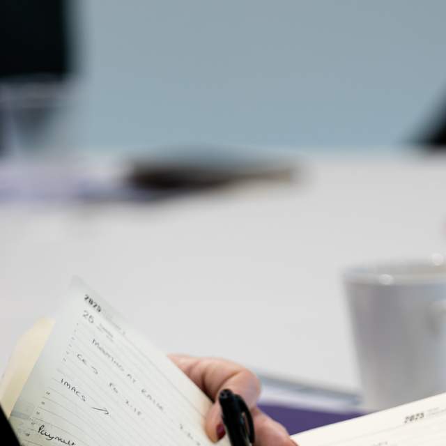 A person writing in a notebook with meeting table and glasses of water in the background.