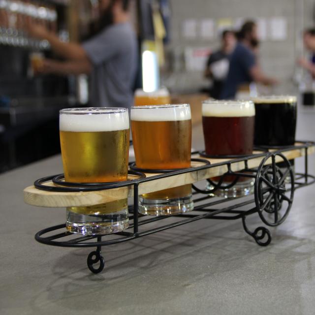 A flight of beer samples displayed apropros in wire-framed bi-plane-shaped holder at Crooked Can Brewing. Flight sits on bar with people in background.