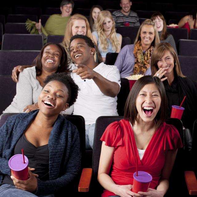 A waist up image of a diverse group of people watching a comedy in a movie theater.