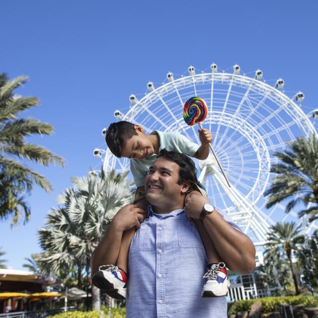 A boy on top of his dad's shoulders, while smiling and holding a lollipop, in front of The Wheel at ICON Park