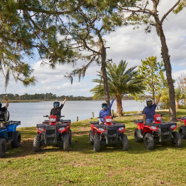 A group of ATV riders raise their fist, as they are parked near the edge of a lake, while underneath the shade of several pine trees at Revolution Adventures.