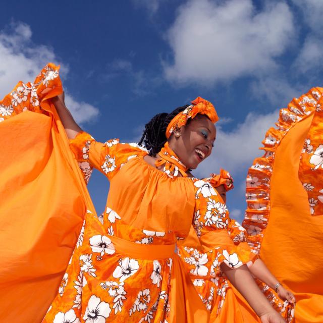 Caribbean dancing troupe wearing bright orange costumes perform on a beach.