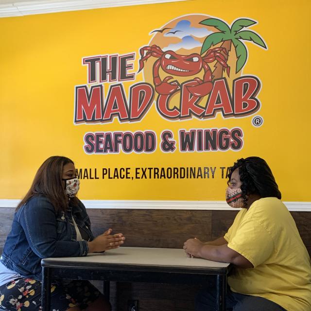 Two women sit facing each other at a table against a bright yellow wall at The Mad Crab Seafood and Wings in Eatonville, Florida.