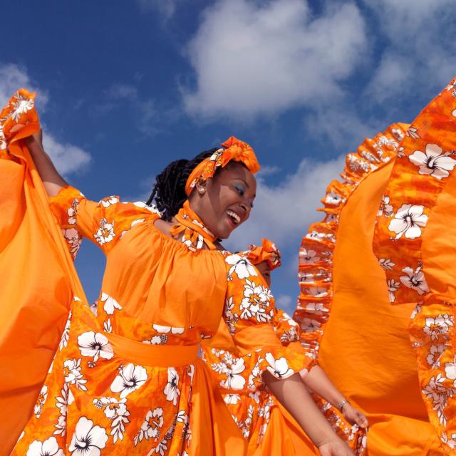 Caribbean dancing troupe wearing bright orange dresses as they perform on a beach.