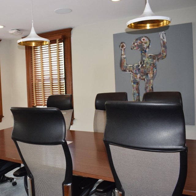 A board room with chairs, a long desk, windows, and art on the wall.