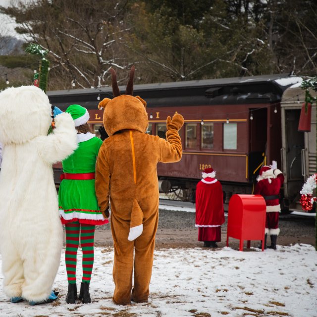 Characters dressed in costumes with Santa and Mrs. Claus waving at passengers on a train.